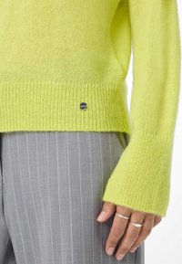 Helle gelbe Strickjacke mit gerippten Bündchen und Saum. Mit einem silbernen Logo-Akzent nahe dem Saum. Kombiniert mit hellgrauen, gestreiften Hosen.