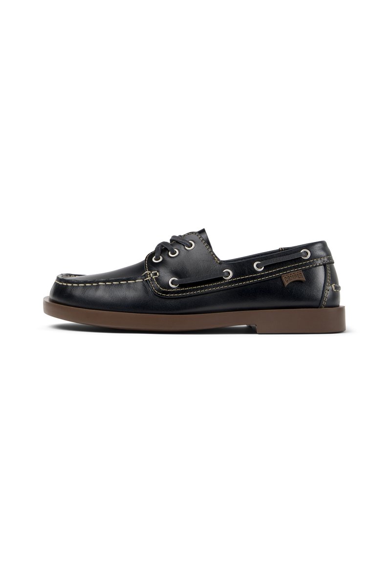 Zapato de barco de cuero negro con suela marrón, costuras beige, puntera redonda, ojales metálicos y un pequeño parche de logo en el lateral.