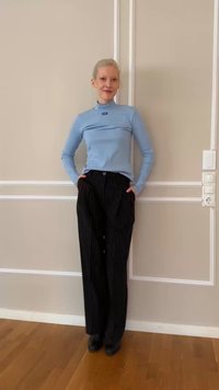 Person standing indoors against beige wall, wearing light blue long-sleeve top and black pinstriped wide-leg pants with hands in pockets.