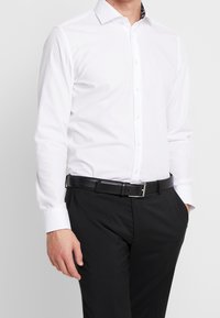 Witte blouse met lange mouwen en een puntige kraag, met een zwart bloemenpatroon aan de binnenkant van de kraag, gecombineerd met zwarte pantalon en een zwarte riem.