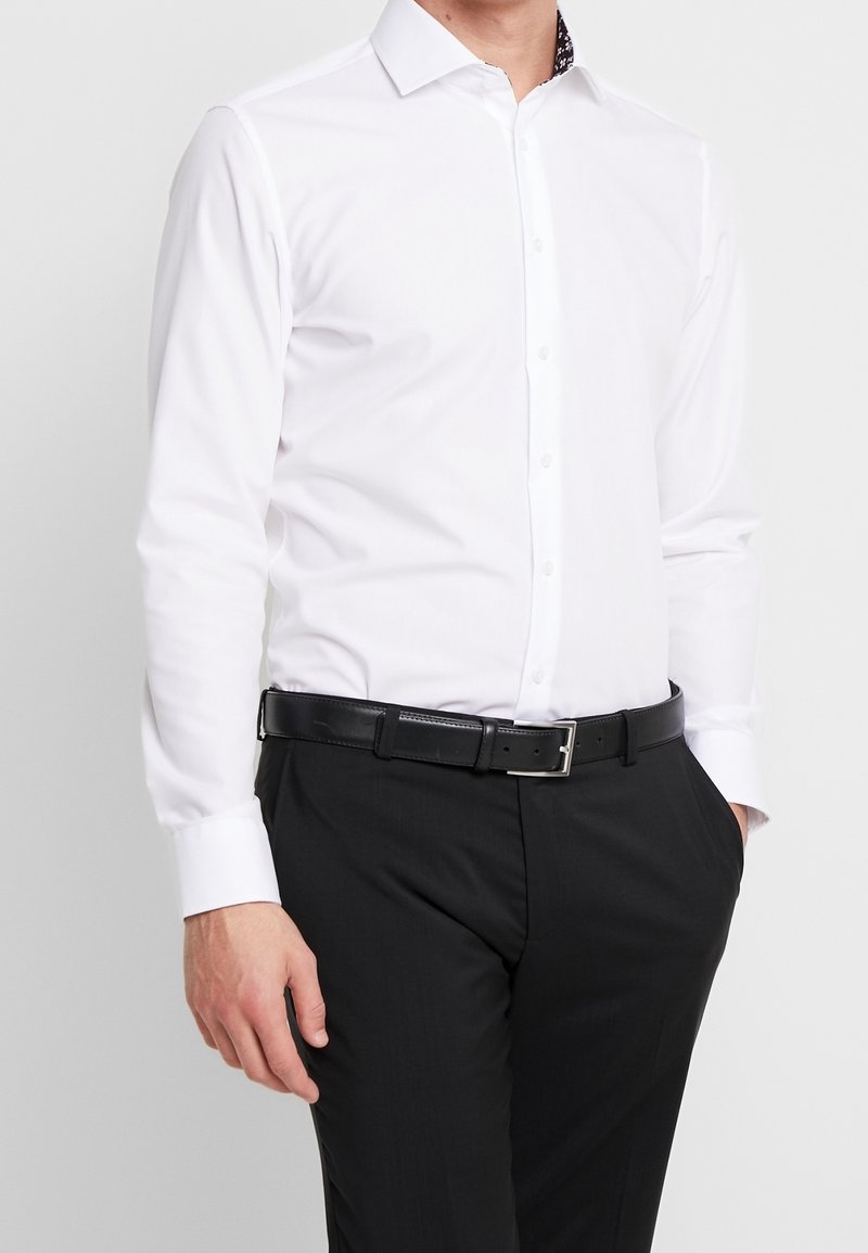Witte blouse met lange mouwen en een puntige kraag, met een zwart bloemenpatroon aan de binnenkant van de kraag, gecombineerd met zwarte pantalon en een zwarte riem.