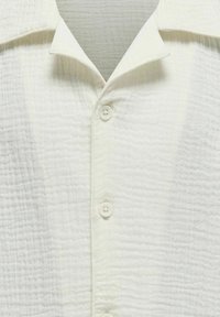 Chemise blanche texturée à manches courtes avec col chemise et boutons sur le devant, présentant un motif discret en grille sur le tissu.