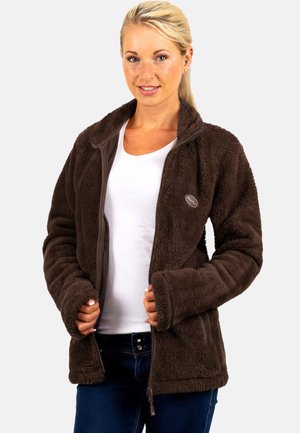 Mujer rubia que lleva una chaqueta de forro polar marrón con cremallera sobre una camisa blanca y vaqueros azules, de pie contra un fondo claro y liso.