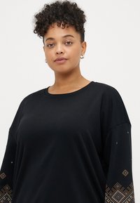 Μαύρη μπλούζα με μακριά μανίκια. Διακοσμητικά σχέδια σε πορτοκαλί και μπλε στα μανίκια. Λείος ύφασμα με στρογγυλή λαιμόκοψη.