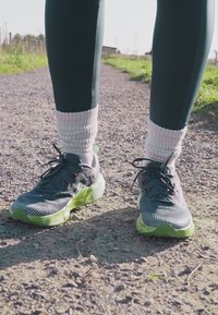 Zapatillas deportivas grises con suelas verdes, parte superior de malla texturizada y acentos rojos. Usadas con calcetines de rayas rosas claros y leggings oscuros.