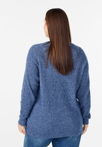 Zils adīts džemperis ar ribotu tekstūru, garām piedurknēm un noapaļotu apakšmalu, kombinēts ar gaiši ziliem džinsiem.