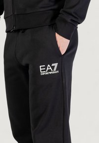 Pantalones de chándal negros de mezcla de algodón, con una cinturilla acanalada, bolsillos laterales y un logo blanco de EA7 Emporio Armani impreso en el muslo.