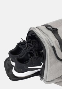 Zaino grigio con un compartimento zip che rivela scarpe da ginnastica nere. La texture è in tessuto e le caratteristiche includono hardware nero e una finitura liscia.