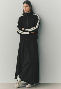 Veste noire zippée avec des rayures blanches, associée à une longue jupe noire et des baskets blanches. Tissu lisse avec des détails de conception athlétique.