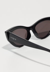 Ochelari de soare negri cu un finisaj lucios, formă supradimensionată de tip cat-eye, lentile închise la culoare și un cadru îndrăzneț, unghiular. Presentă branding-ul "vogue" pe lateral.