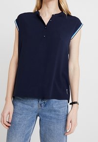 Top però a maniche corte di colore blu navy con colletto, caratterizzato da due bottoni frontali e dettagli blu a contrasto sulle maniche. Tessuto leggero e vestibilità rilassata.