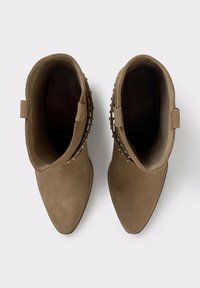 Botas de tobillo de ante marrón con punteras puntiagudas, detalles de tachuelas a los lados y lengüetas en los talones. Forro interior texturizado.