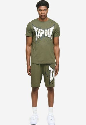 Młody mężczyzna stoi, ubrany w oliwkowozielone szorty i pasującą koszulkę z dużym nadrukiem "TAPOUT", białe skarpetki i białe trampki.