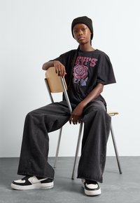 Camiseta gráfica negra con diseño de rosa, jeans negros de corte amplio, zapatillas negras con acentos blancos, sentado en una silla de metal y madera.