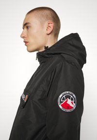 Giacchetto nero con cappuccio e zip sul davanti, caratterizzato da un logo sul petto e una toppa rotonda sulla manica. Tessuto liscio e leggero.