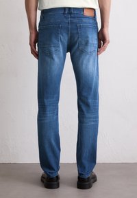 Jean en denim bleu slim fit avec un délavage, poches arrière et une étiquette en cuir sur la taille. Présente un léger effet usé sur les cuisses.