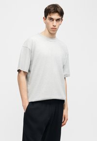 Νέος άνδρας που φοράει μια ανοιχτόγριζη φαρδιά μπλούζα και μαύρο παντελόνι, στέκεται με το ένα χέρι στην τσέπη μπροστά σε απλό φόντο.