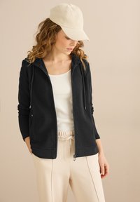 Sudadera con cremallera negra de tejido texturizado, con capucha ajustable y bolsillos laterales, combinada con una camiseta blanca y pantalones beige, rematada con un sombrero crema.