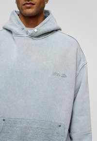 Lichtblauwe oversized hoodie met een zachte textuur, voorzien van een textuurkap, een frontzak en subtiele geborduurde logo op de borst.