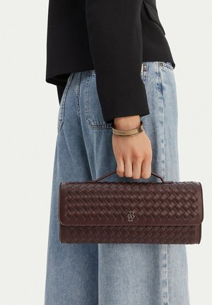 Osoba držící tmavě hnědou pletenou koženou kabelku za horní rukojeť, oblečená v světle modrých džínách a černém topu s dlouhým rukávem.