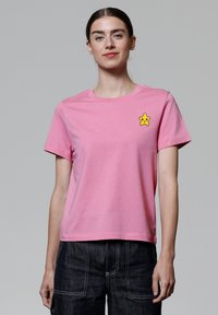 Pinke Baumwoll-T-Shirt mit Rundhalsausschnitt und kurzen Ärmeln, versehen mit einem gelben Stern-Patch auf der linken Brust.