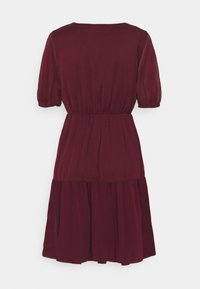 Burgunderfarbenes Kurzarmkleid mit elastischem Bund, geraffter Naht unterhalb der Hüften und einem gestuften Rock vor einem weißen Hintergrund.