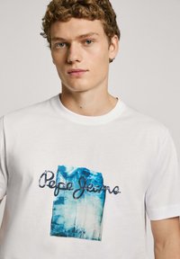 Balta marškinėliai su mėlynu grafiniu spaudiniu, ant kurio užrašas "Pepe Jeans" juoda spalva, esantis ant papilkėjusio mėlyno fono. Trumpomis rankovėmis.