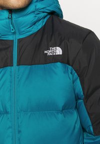 Veste matelassée bleu sarcelle avec panneaux noirs. Comprend une capuche, une fermeture éclair à l'avant et un logo sur la poitrine. Tissu texturé avec un motif en grille.