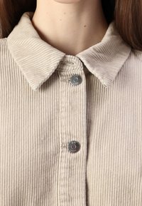 Beige kordfløyelskjorte med kragehals, med to metalltrykkknapper og vertikal ribbestruktur gjennom hele.