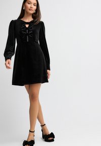 Schwarzes Samtkleid mit gepufften Ärmeln, einer Bogenverzierung am Hals und einem ausgestellten Rock. Kombiniert mit schwarzen Blockabsätzen, verziert mit Schleifen.