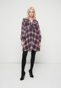 Κοντό φόρεμα σε μπεζ και γκρι καρό σχέδιο με φρου φρου στο λαιμό, μακριά μανίκια και φουσκωτό φουστάνι, συνδυασμένο με μαύρες κολάν και ψηλοτάκουνα παπούτσια.