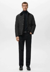 Giacca trapuntata nera con colletto morbido, abbinata a un dolcevita nero, pantaloni scuri e scarpe nere lucide. Design minimalista.