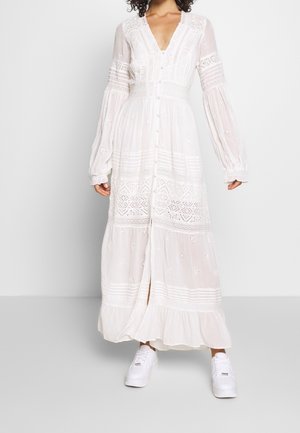 Femme portant une longue robe en dentelle blanche avec des boutons sur le devant et des baskets blanches, debout devant un fond clair uni.