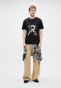 T-shirt de algodão preta com um gráfico de um cão na parte da frente, combinada com calças largas beiges e uma flanela xadrez amarrada na cintura.