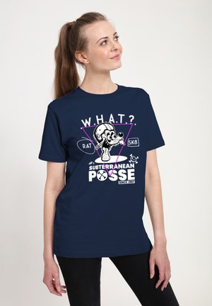 Ung kvinde med hestehale iført navyblå T-shirt med tegneserie rotte med hjelm og teksten "W.H.A.T? Subterranean Posse Since 1967."