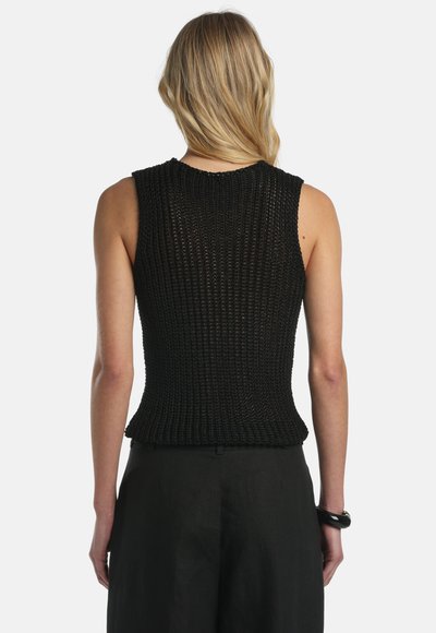 Ženska z blond lasmi, oblečena v brezrokavnik iz črne pletene tkanine in črne hlače, prikazana od zadaj na enostavni podlagi.