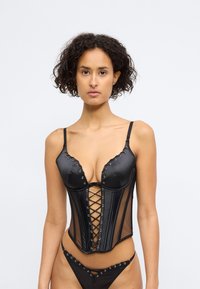 Cuerpo negro con escote en V, frente con cordones y paneles de malla. Las correas ajustables y los detalles de hardware en contraste realzan el diseño.
