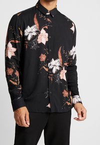 Chemise à manches longues noire avec un motif floral en rose et blanc, présentant un design à boutons et une texture de tissu douce.