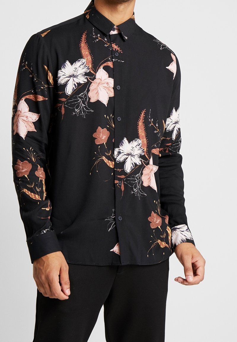 Chemise à manches longues noire avec un motif floral en rose et blanc, présentant un design à boutons et une texture de tissu douce.