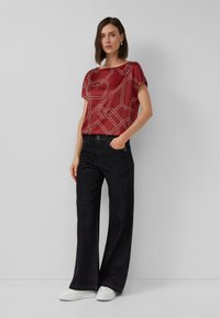 Bordeauxfarbenes kurzärmliges Oberteil mit weißem abstraktem Muster, kombiniert mit dunklen weiten Jeans und weißen Sneakers, vor einem neutralen Hintergrund.