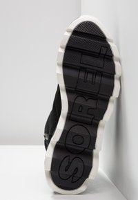 Semelle d'une sneaker noire avec de larges bords blancs, un gros motif de semelle, et le mot "SOREL" en relief au fond.