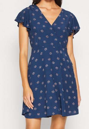 Vrouw draagt een marineblauwe jurk met korte mouwen, kleine roze en witte bloemenpatronen en een V-hals.
