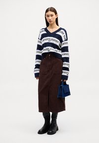 Donna con un maglione a righe blu navy e bianche di Tommy Jeans, una gonna midi marrone con spacco frontale, stivali neri e una piccola borsa blu.