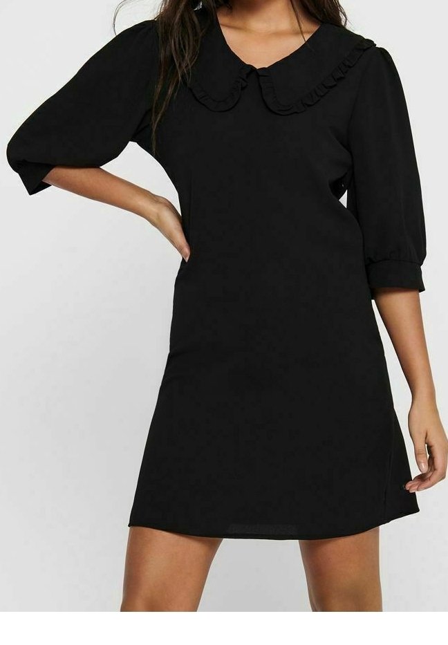 Femme portant une robe noire avec des manches trois-quarts bouffantes et un grand col claudine volanté, debout une main sur la hanche.