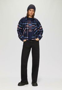Geverfde flanellen jas in marineblauw en roze, zwart graphic T-shirt, zwarte wijde jeans en zwarte platform schoenen; gebreide beanie op het hoofd.