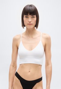 Γυναίκα με κοντά καστανά μαλλιά που φοράει ένα λευκό ριμπ λευκό σουτιέν και μαύρο σλιπ υψηλής κοπής, στέκεται μπροστά σε απλό φωτεινό φόντο.