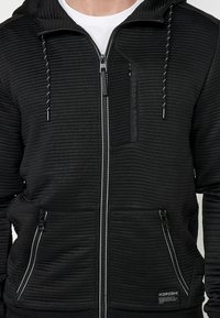 Felpa nera a costine con cerniera frontale, tasche anteriori, cerniera verticale sul petto, cursori con marchio e camicia bianca visibile sotto.