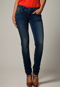 Kvinna i mörkblå skinny jeans, röd topp och bruna högklackade skor, med en hand i fickan och den andra handen avslappnad.