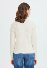 Femme aux cheveux bruns mi-longs portant un pull en tricot crème et un jean bleu, tournant le dos contre un fond uni.