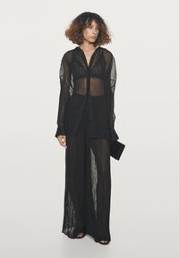 Black sheer long-sleeve shirt with button front, layered over a matching black pleated wide-leg pant, featuring delicate fabric texture.